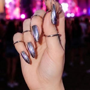 May include: A hand adorned with multiple thin, plain metal rings on the fingers and a delicate chain bracelet on the wrist. The fingernails are long, pointed, and finished with a highly reflective, smooth, metallic silver chrome polish.