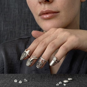 May include: A set of four silver-toned nail rings worn on the fingers. The rings feature intricate metalwork, including textured surfaces, small embedded stones, and floral-inspired patterns that extend over the nail bed area.