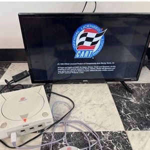 May include: A white Sega Dreamcast video game console with a controller connected by a grey cord, positioned in front of an Onn television screen displaying a black background with the Official Licensed Product CART logo and copyright text.