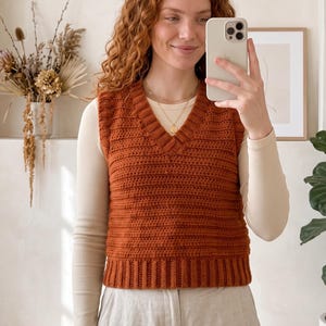 May include: A rust-coloured sleeveless crochet sweater vest featuring a V-neckline, a ribbed hem, and a horizontal textured stitch pattern. The garment is worn over a long-sleeved cream-coloured top, creating a layered look with a classic silhouette.