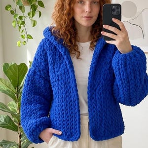 May include: A vibrant royal blue chunky knit cardigan featuring a hood, long sleeves, and a thick, textured yarn construction. The garment is worn over a plain white t-shirt and light-colored trousers, showcasing a relaxed fit and open front.