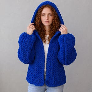 Può includere: Un cardigan in maglia grossa di colore blu reale vibrante con cappuccio e maniche lunghe. Il capo è realizzato in filato spesso e testurizzato con un design aperto sul davanti, che mette in risalto un motivo a maglia pesante e denso.