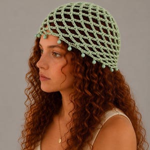 May include: A light sage green crochet beanie featuring an open diamond-shaped mesh pattern. The lower edge of the cap is finished with a row of small, matching sage green spherical beads dangling from each point of the crochet netting.