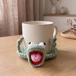 May include: A light green crocheted mug cosy featuring a frog-like face with white and black eyes, a wide open mouth with a red interior, and a textured pink tongue. The cosy wraps around a plain off-white ceramic mug, providing a decorative cover.