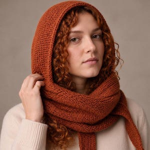 May include: A rust-coloured knitted hooded scarf featuring a textured stitch pattern. The accessory consists of a hood that transitions into a long scarf, crafted from a solid-coloured yarn. The design provides full head and neck coverage.