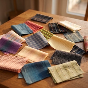 May include: A collection of rectangular fabric swatches in various colors and patterns, including plaid, checkered, and solid weaves in shades of blue, red, yellow, green, and grey, arranged in a scattered layout on a wooden surface.