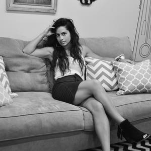 May include: A woman sits on a gray couch with pillows. She is wearing a white tank top and a black skirt. She has long dark hair and is looking at the camera.