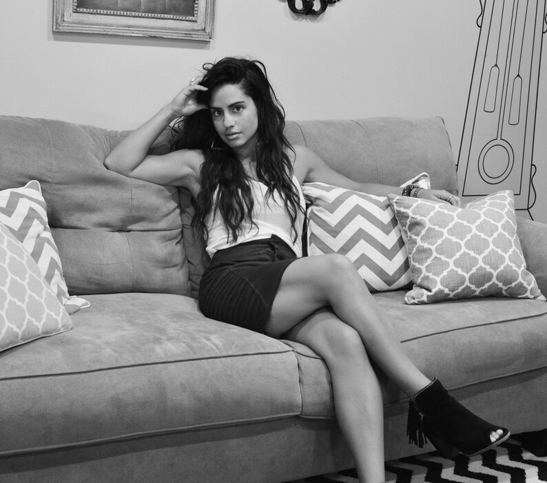 May include: A woman sits on a gray couch with pillows. She is wearing a white tank top and a black skirt. She has long dark hair and is looking at the camera.