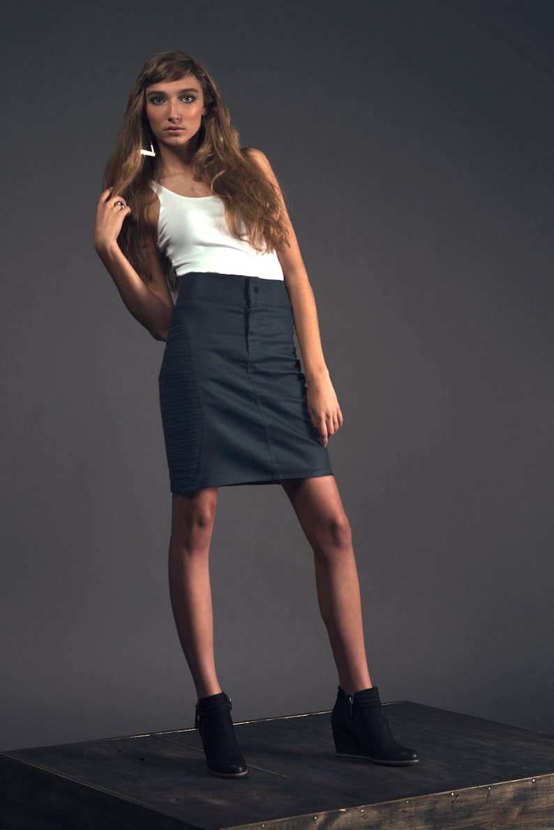 May include: A woman wearing a white tank top and a dark gray pencil skirt stands on a wooden platform. She is wearing black ankle boots with a wedge heel.