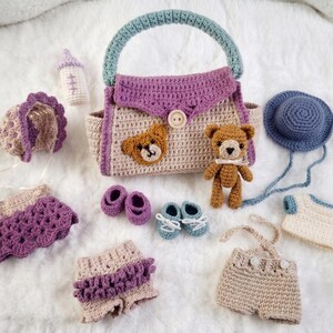 Peut inclure: Un ensemble au crochet comprenant un sac beige avec une anse bleu sarcelle et un rabat violet, un petit ours en peluche marron, un bonnet violet, une jupe violette, un short beige à volants violets, une salopette beige, un chapeau bleu et deux paires de petits chaussons.