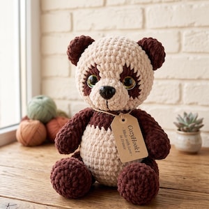 Peut inclure: Une peluche panda crochetée en fil épais et doux de couleur crème et marron foncé. Elle présente de grands yeux sombres brillants, un nez en plastique noir et des oreilles rondes. Une étiquette en carton beige porte le texte CozyHook1 Made with Love in France.