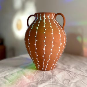 Könnte beinhalten: Eine terrakottafarbene Keramikvase mit zwei Henkeln, die mit einem Muster aus vertikalen weißen Linien und kleinen weißen Punkten verziert ist. Die Vase hat einen bauchigen Körper und eine weite Öffnung mit einer matten Außenoberfläche.