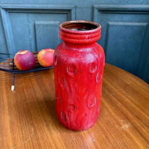 Puede incluir: Un jarrón cilíndrico de cerámica con un esmalte rojo vibrante y una superficie texturizada de hendiduras circulares en relieve. El jarrón tiene un cuello estrecho con un borde grueso y redondeado y un acabado interior de color marrón oscuro.