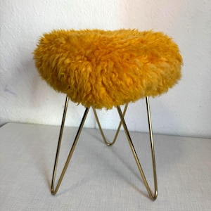 May include: A small round stool featuring a thick, textured mustard yellow faux fur seat cushion. The seat is supported by four thin, gold-toned metal hairpin legs that angle outward from the center to provide a stable base for the furniture piece.
