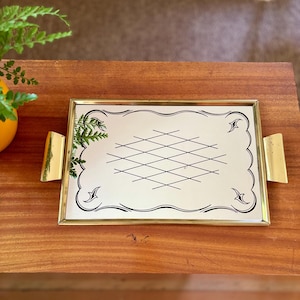 Op de afbeelding: Een rechthoekig dienblad met een spiegelend oppervlak, een zwart ruitpatroon en decoratieve hoekaccenten. Het dienblad is omlijst met gepolijst goudkleurig metaal en heeft twee bijpassende goudkleurige metalen handgrepen aan de zijkanten.
