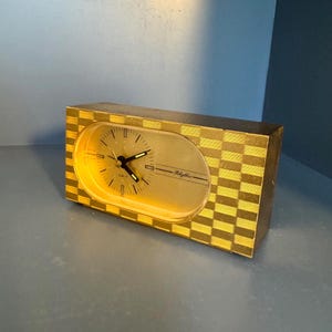 May include: A rectangular quartz clock with a gold and dark brown checkered pattern on the exterior. The clock face is oval-shaped with a gold background, black hands, and the word Rhythm printed in black script on the right side of the dial.