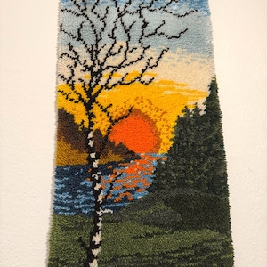 Könnte beinhalten: Ein rechteckiger Punch-Needle-Wandbehang mit einer Landschaftsszene, die einen hohen, kahlen Baum im Vordergrund, eine leuchtend orangefarbene Sonne über einem blauen See und dunkelgrüne Kiefern vor einem hellblauen und gelben Himmel zeigt.