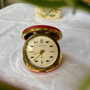 Könnte beinhalten: Ein vintage Europa Reise-Wecker mit einem roten, ledertexturierten Klappgehäuse und einem goldfarbenen Metallrahmen. Das runde Zifferblatt ist cremefarben mit Gittermuster, goldenen Zeigern, schwarzen Stundenmarkierungen und der Aufschrift Europa 2 Jewels Germany.
