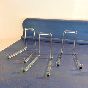 Op de afbeelding: Drie doorzichtige acryl displaystandaards, elk met een L-vormig ontwerp met een verticale rugsteun en een horizontale basis. De standaards zijn gemaakt van transparant materiaal en hebben kleine, afgeronde uiteinden aan de basis.