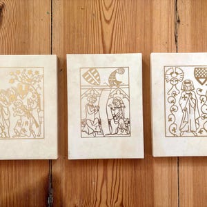 May include: Three rectangular off-white plaques featuring gold-colored line art illustrations. Each plaque displays a distinct medieval-style scene with figures, heraldic shields, and foliage patterns, all enclosed within a thin gold rectangular border.
