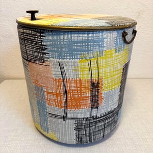 Pode incluir: Um balde de gelo cilíndrico de metal com um padrão de hachuras cruzadas multicoloridas em amarelo, laranja, azul claro e preto. O balde inclui uma tampa com um pequeno puxador preto e uma argola lateral de metal para transporte.