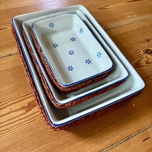 Könnte beinhalten: Ein Set aus drei ineinander stapelbaren rechteckigen Keramik-Auflaufformen mit weißem Innenraum und blauen Blumenmustern. Jede Form hat einen blauen Rand und ist in einen geflochtenen braunen Weidenkorb eingefasst, was dem Set ein geschichtetes Aussehen verleiht.