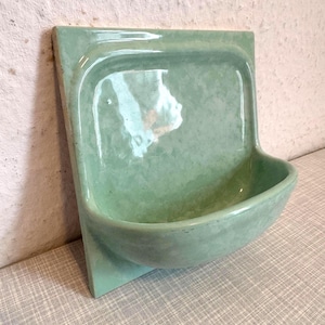 Può includere: Un portasapone da parete in ceramica con una finitura lucida color verde menta chiaro. L'oggetto presenta una piastra posteriore rettangolare piatta e un bacino semicircolare con un interno liscio e curvo, ideale per riporre piccoli oggetti.