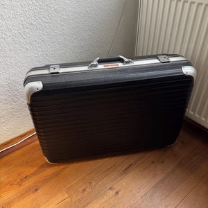 Puede incluir: Una maleta rectangular rígida de color negro con detalles acanalados horizontales, refuerzos de metal plateado en las esquinas, un asa central de transporte y dos cierres de seguridad. La maleta tiene un perfil delgado y acabado oscuro.