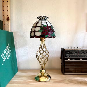 Può includere: Una lampada da tavolo in vetro colorato con un paralume a cupola decorato con motivi floreali bianchi, rossi e verdi. La lampada presenta una base in metallo dorato lucido con uno stelo a filo intrecciato a spirale e un supporto circolare.