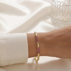 Op de afbeelding: Een gouden armband met twee rechthoekige segmenten ingelegd met kleine, levendige roze steentjes. De gepolijste gouden band heeft een glad, afgerond profiel en de roze stenen accenten zijn verzonken in het metalen oppervlak van de armband.