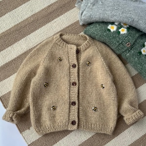 Könnte beinhalten: Eine beigefarbene Strickjacke mit langen Ärmeln, Rundhalsausschnitt und Knopfleiste auf der Vorderseite. Das Kleidungsstück ist mit kleinen, gelb-schwarz bestickten Bienenmotiven verziert, die über die Vorderseite und Ärmel verteilt sind.