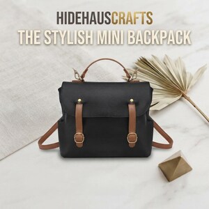 Könnte beinhalten: Ein schwarzer Mini-Rucksack mit hellbraunen Riemen, goldfarbenen Schnallen und einem Tragegriff. Der Text HIDEHAUSCRAFTS THE STYLISH MINI BACKPACK steht über der Tasche. Die Tasche hat einen Klappverschluss mit zwei vertikalen Riemen und Metallbeschlägen.