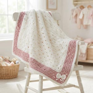 May include: A cream-colored crocheted baby blanket featuring a textured shell stitch pattern and a wide dusty rose border. The blanket is accented with a small, white crocheted bow detail at the bottom corner, draped over a white wooden rocking chair.