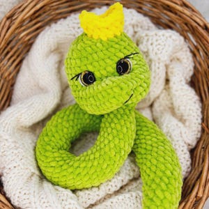 Peut inclure: Un serpent en peluche crocheté vert citron avec une couronne jaune vif sur la tête. Le serpent possède de grands yeux ronds de sécurité noirs avec des reflets dorés et des cils brodés. La texture est douce et moelleuse, avec un corps enroulé.