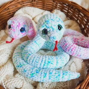 May include: Two plush snakes made of soft, variegated yarn in shades of pastel pink, blue, and white. Each snake features large, glossy dark eyes and a small, bright red felt tongue protruding from its mouth. The bodies are coiled and textured.