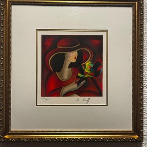 May include: A framed print featuring a stylized illustration of a figure wearing a wide-brimmed red hat and red clothing, holding a colorful bouquet. The artwork is signed Le Kinff and numbered 325/350, set within a decorative gold-toned frame.