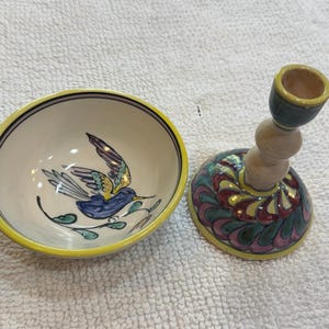 Może przedstawiać: Ceramiczna miska i pasujący świecznik. Miska ozdobiona jest ręcznie malowanym niebieskim ptakiem z żółtymi i fioletowymi skrzydłami na kremowym tle z żółtym brzegiem. Świecznik ma okrągłą podstawę z wirującym fioletowo-zielono-żółtym wzorem oraz wysoki, jasny trzon.