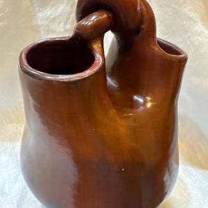 May include: A polished, reddish-brown ceramic vessel featuring two distinct openings connected by a central, arched handle. The surface has a smooth, glossy finish with subtle variations in color, showcasing a rounded, bulbous base and a sculptural form.