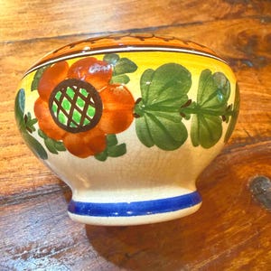 Könnte beinhalten: Eine Keramikschale mit cremefarbener Basis und einer rissigen Glasuroberfläche. Die Außenseite ist mit handgemalten orangefarbenen Blumen mit grünen Zentren und grünen Blättern verziert, akzentuiert durch ein gelbes Band und einen blauen Rand.