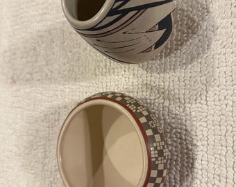 Puede incluir: Dos cuencos de cerámica con formas redondeadas y aberturas superiores. El cuenco superior presenta una base crema con patrones geométricos negros. El cuenco inferior tiene un interior crema y un exterior decorado con una cuadrícula de pequeños cuadrados negros y blancos.