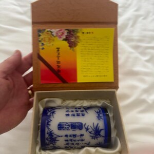 May include: A cylindrical white ceramic vessel with blue bamboo illustrations and black Chinese calligraphy, resting in a beige fabric-lined wooden box. The box lid features a yellow informational card with floral imagery and printed text.
