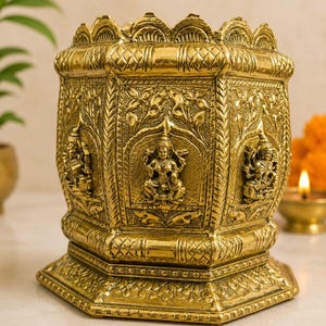 May include: A gold-colored brass vessel with an octagonal shape, featuring intricate embossed relief carvings of seated deities on each panel. The top rim has a scalloped edge, and the base is wide with detailed geometric and floral patterns.