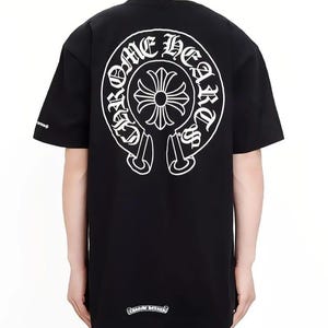 May include: A black short-sleeve t-shirt featuring a large white circular graphic on the back with the text Chrome Hearts and a central cross design. A smaller white cross logo is printed on the right sleeve and near the bottom hem.