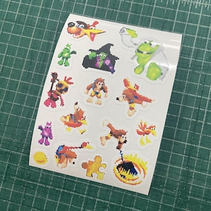 Op de afbeelding: Een wit stickervel met diverse pixel art personages en iconen uit de Banjo-Kazooie videogame-serie, waaronder Banjo, Kazooie, Gruntilda, Mumbo Jumbo en een gouden puzzelstukje, allemaal gerangschikt op een enkele zelfklevende achterkant.
