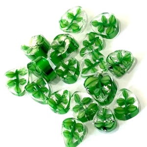Puede incluir: Una colección de cuentas de vidrio transparente con diseños de hojas verdes. Las cuentas son de varias formas y tamaños, con patrones de hojas verdes visibles dentro del vidrio transparente. Las cuentas están dispersas sobre una superficie blanca.