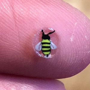 May include: A tiny, detailed bee design with black and yellow stripes, white wings, and black antennae. The bee is set within a clear, circular shape. The image is a close-up, showcasing the intricate details of the miniature artwork.