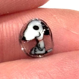 Puede incluir: Un amuleto de vidrio transparente con una ilustración en blanco y negro de Snoopy, un beagle de dibujos animados, en su interior.