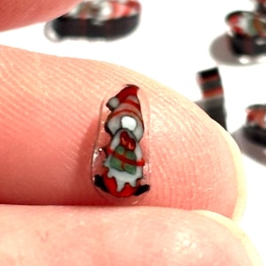 Puede incluir: Una pequeña decoración de arte de uñas de gnomo navideño. El gnomo lleva un gorro a rayas rojas y blancas y un abrigo rojo. El gnomo tiene una barba blanca y un regalo verde y rojo. El arte de uñas está sobre un fondo transparente.