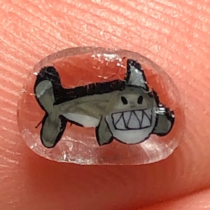 Può includere: Un design per nail art trasparente e ovale con uno squalo dei cartoni animati. Lo squalo è dipinto in tonalità di grigio, bianco e nero, con un ampio sorriso e denti affilati. Il design è applicato su un'unghia.