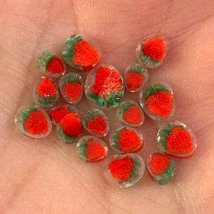Puede incluir: Una colección de pequeñas cuentas de vidrio con forma de fresa. Cada cuenta presenta un diseño de fresa rojo vibrante con hojas verdes, encapsulado en vidrio transparente. Las cuentas varían en tamaño y están dispuestas sobre una superficie clara.